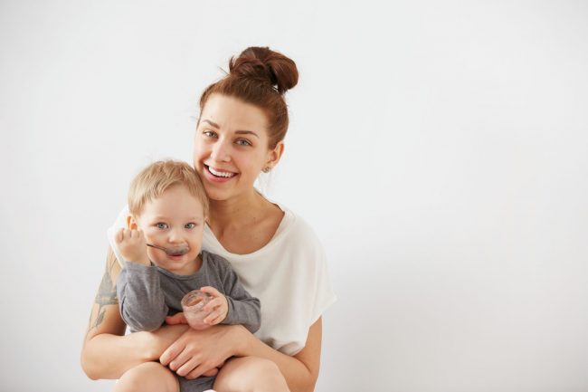 Young attractive mother with her one years old little son dressed in pajamas. Boy eating a fruit smoothie himself in the bedroom at the weekend together, warm and cozy scene. Selective focus.