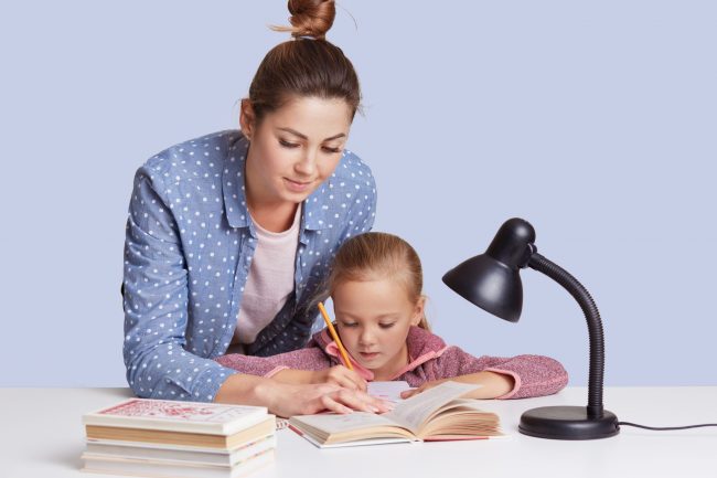 Beautiful Caucasian woman helping her doughter to do school homework, mother and child surronded by books, little girl sitting concentrated at white desk, trying to do sums. Education concept.